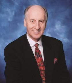 Smiling man with thinning hair wearing a black suit and patterned tie against blue.
